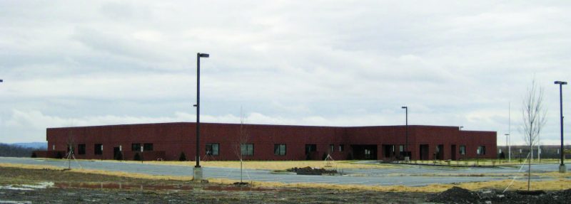 A large red brick one-story building under a cloudy sky and behind an empty parking lot with three tall street lamps.