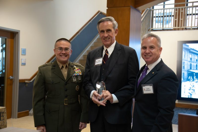 Three well dressed men stand next to each other, with the man in the middle holding a shiny glass award. They are facing the camera all smiling.