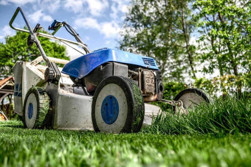 A blue lawnmower sits motionless on a patch of lush grass while blades of grass stick to the mower body.