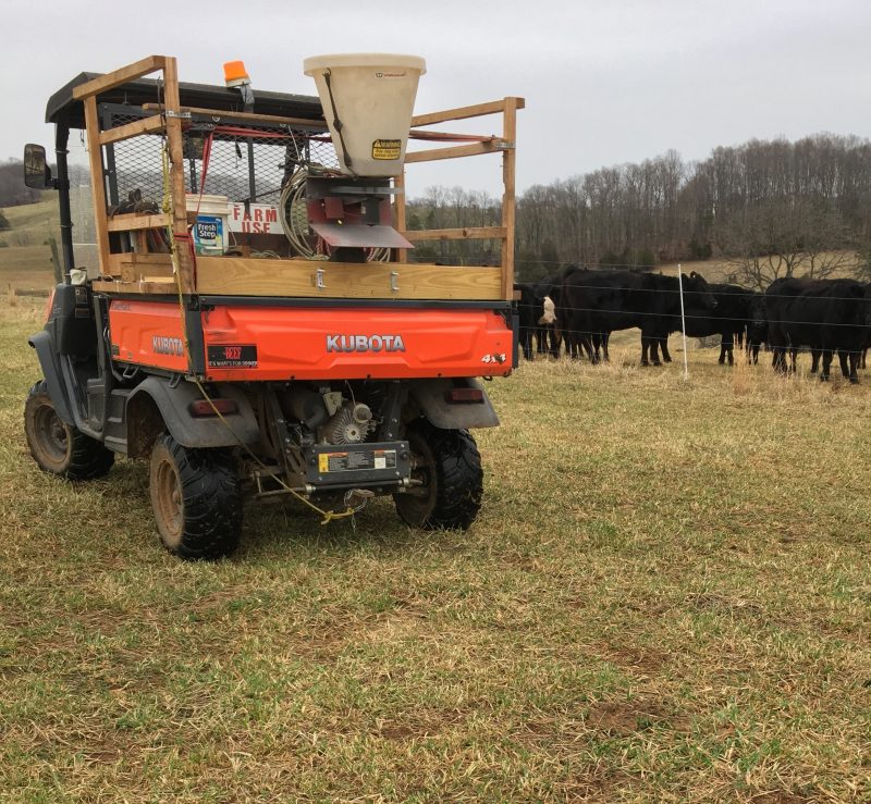 A Kubota utility vehicle with a seed spreader parked in a field, broadcasting clover seed in late winter; black cows graze behind an electric fence with trees and hills in the background.