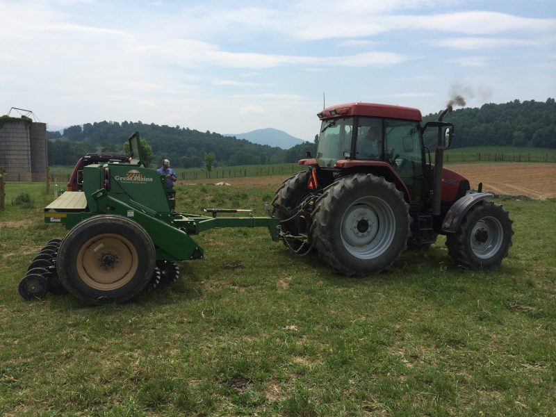 A red tractor pulling a green no-till drill in a field, with a person standing nearby; rolling hills, trees, and a silo are visible in the background.