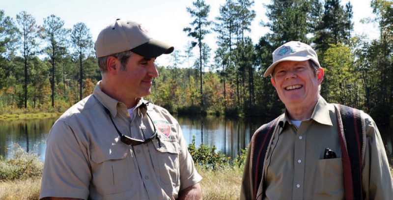 A close-up view of a Virginia Department of Forestry forester standing next to and smiling at a laughing older landowner. They are outdoors and standing in front of a small pond that is surrounded by pine trees. 