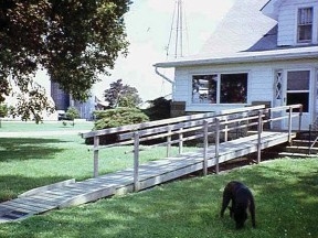 An image of a white house in a grassy yard with a large tree with a long, wooden handicap ramp set on a grassy yard.