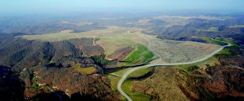 Aerial view depicting flat brown land in between mountains with a patch of green land in the very center and a road traversing the site.