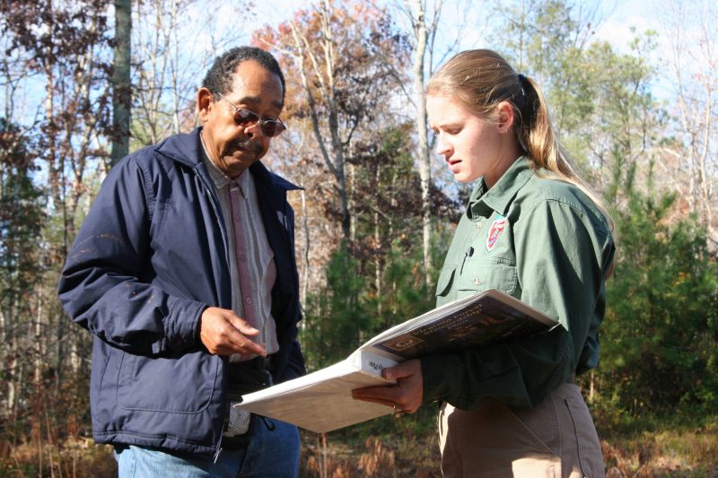 A female forester in green uniform speaks with a man dressed in blue. They stand among tall trees, possibly discussing forest health or management practices as they refer to a book the forester is holding.