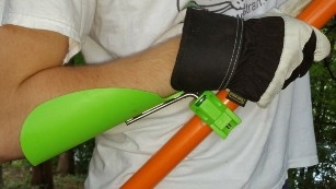 An orange handle being held by a gloved hand. The arm is supported by a long-handled tool leverage booster.