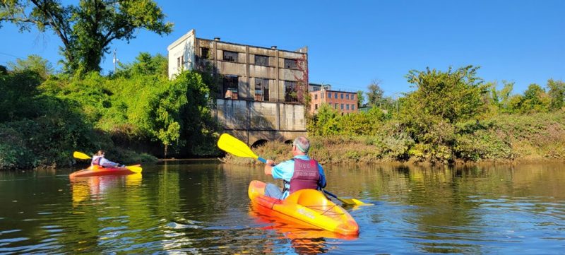 Two people in brightly colored kayaks on a river in front of a large building on the riverbank.