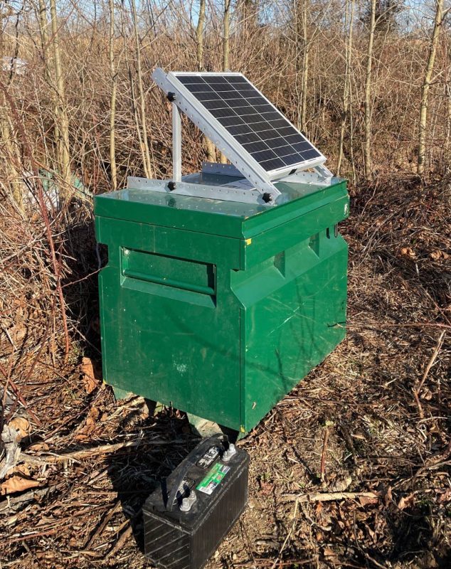 A green rectangular water monitoring unit equipped with a slanted solar panel, placed in a forested area. A black battery box lies on the ground next to the unit.