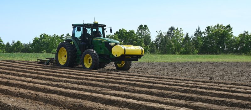 green tractor in brown rows of soil under a blue sky and in front of green trees