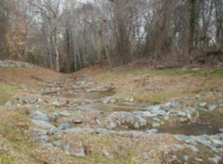 A grassy area surrounded by woods with a path of three roughly circular shallow pools, each dammed with rock and dirt, allowing water to flow gently down an incline. 