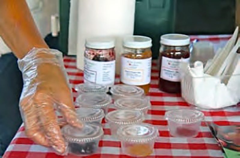 clear cups with clear lids on a red gingham tablecloth and in front of jars of the produce and a clear gloved hand placing one of the samples.