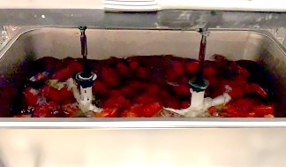 A metal pan two-thirds filled with strawberries in water with two pads stirring the water.