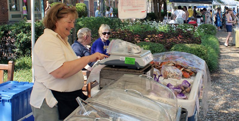 A seafood vendor standing behind a display of covered products uses a scale to weigh a bag containing seafood. Other vendor tents and customers are in the background.
