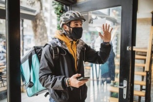Male delivery person wearing mask with bike helmet, thermal bag carring food, cell phone in one hand, and waving to signal arrival of food with other hand. 