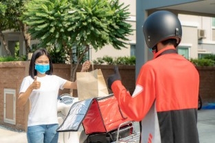 Asian woman pick up delivery food bag from box and thumb up form contactless or contact free from delivery rider with bicycle in front house for social distancing for infection risk.