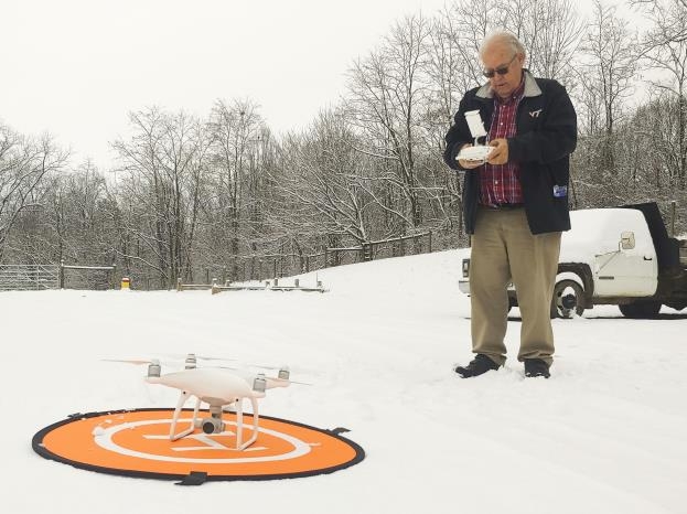 A drone sitting on top of a nylon orange circular landing pad lying on snow-covered ground with a man standing behind it holding the controller.