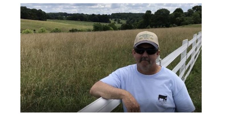Man standing near a white fence wil a field of tall fescue in background.