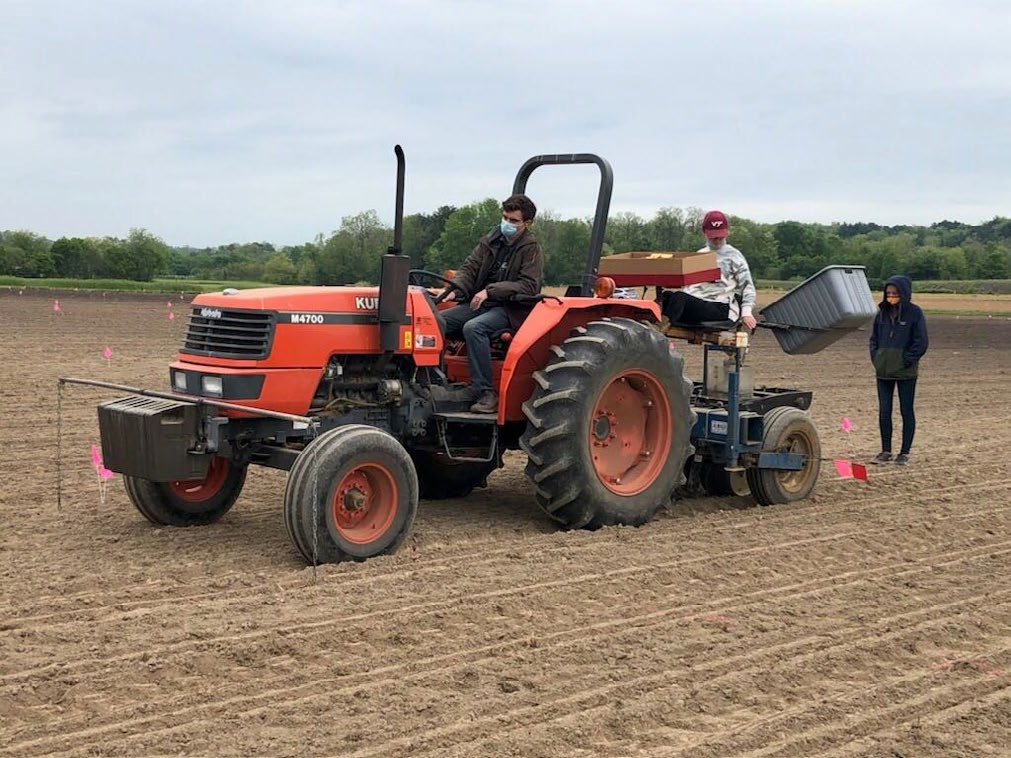 A man drives a planter over dry, bare ground with a helper on the back of the planter and a woman observes from behind.